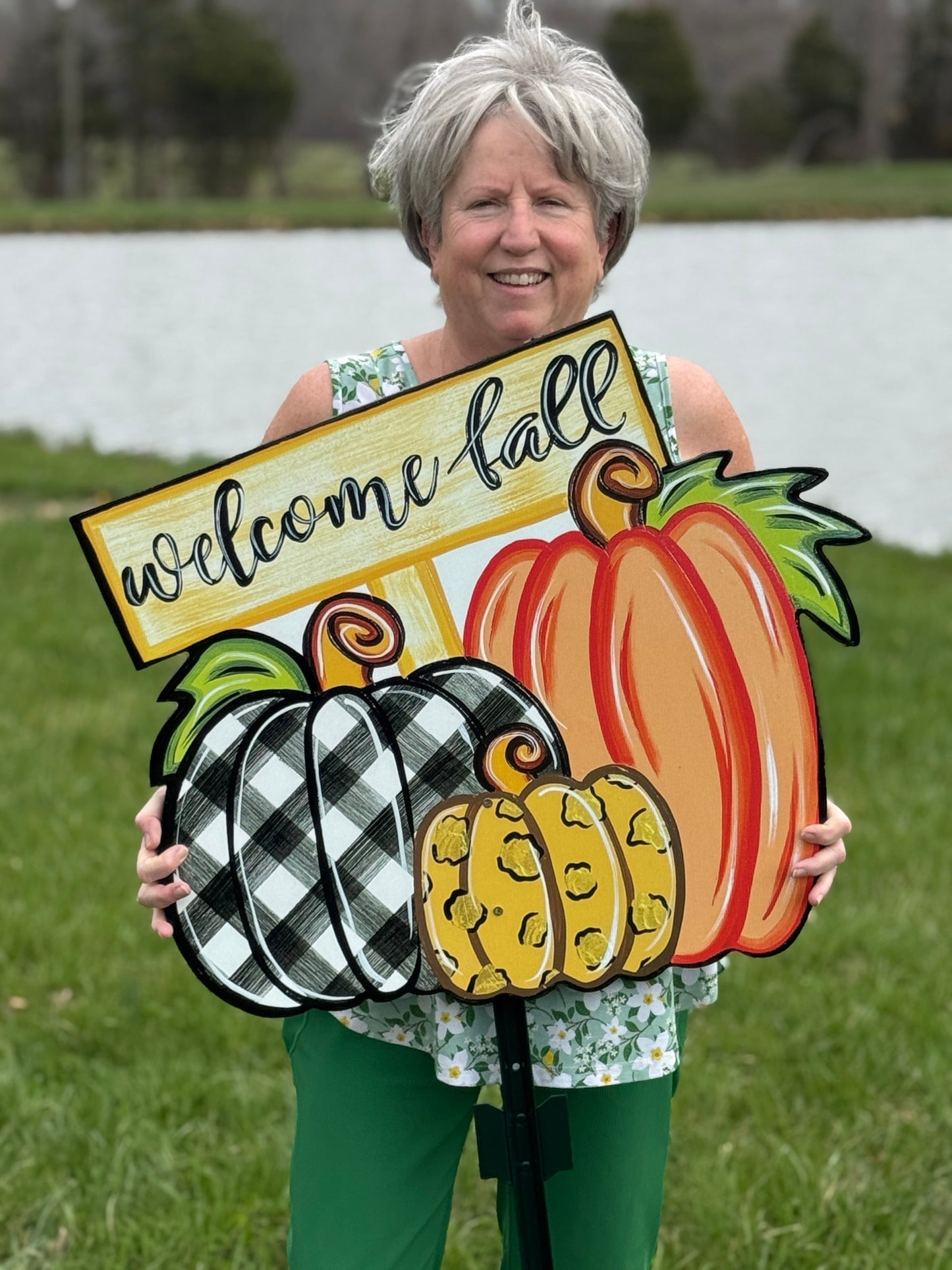 person holding Welcome Fall Sign on top of three Fall pumpkins yard art decoration: one pumpkin is black and white plaid, one pumpkin is leopard print, and one pumpkin is orange