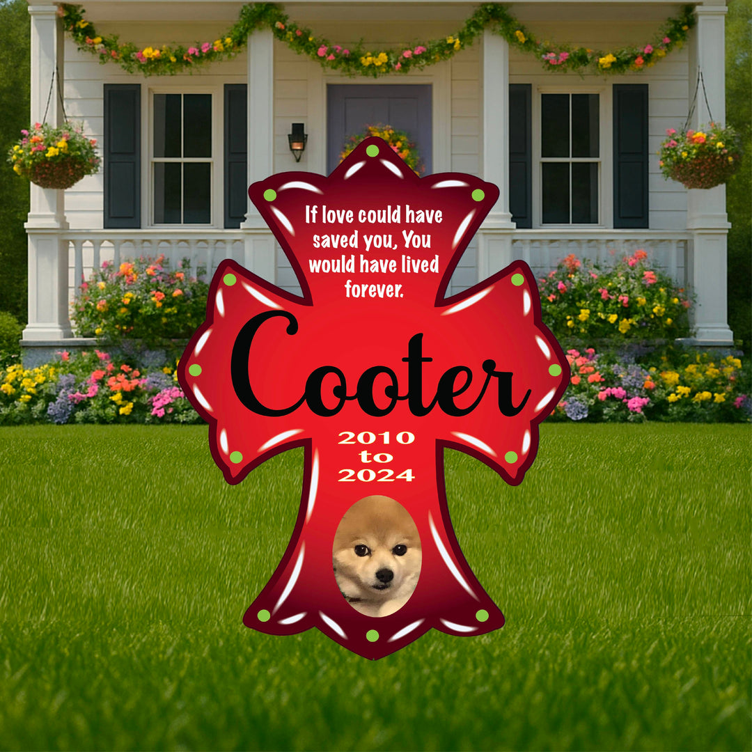 Red cross-shaped pet memorial plaque with white text that reads "If love could have saved you, You would have lived forever." The name "Cooter" is written in large script, along with the years "2010 to 2024." At the bottom is a photo of a small tan dog’s face. The plaque is displayed on a grassy lawn in front of a house with a flower-filled garden.