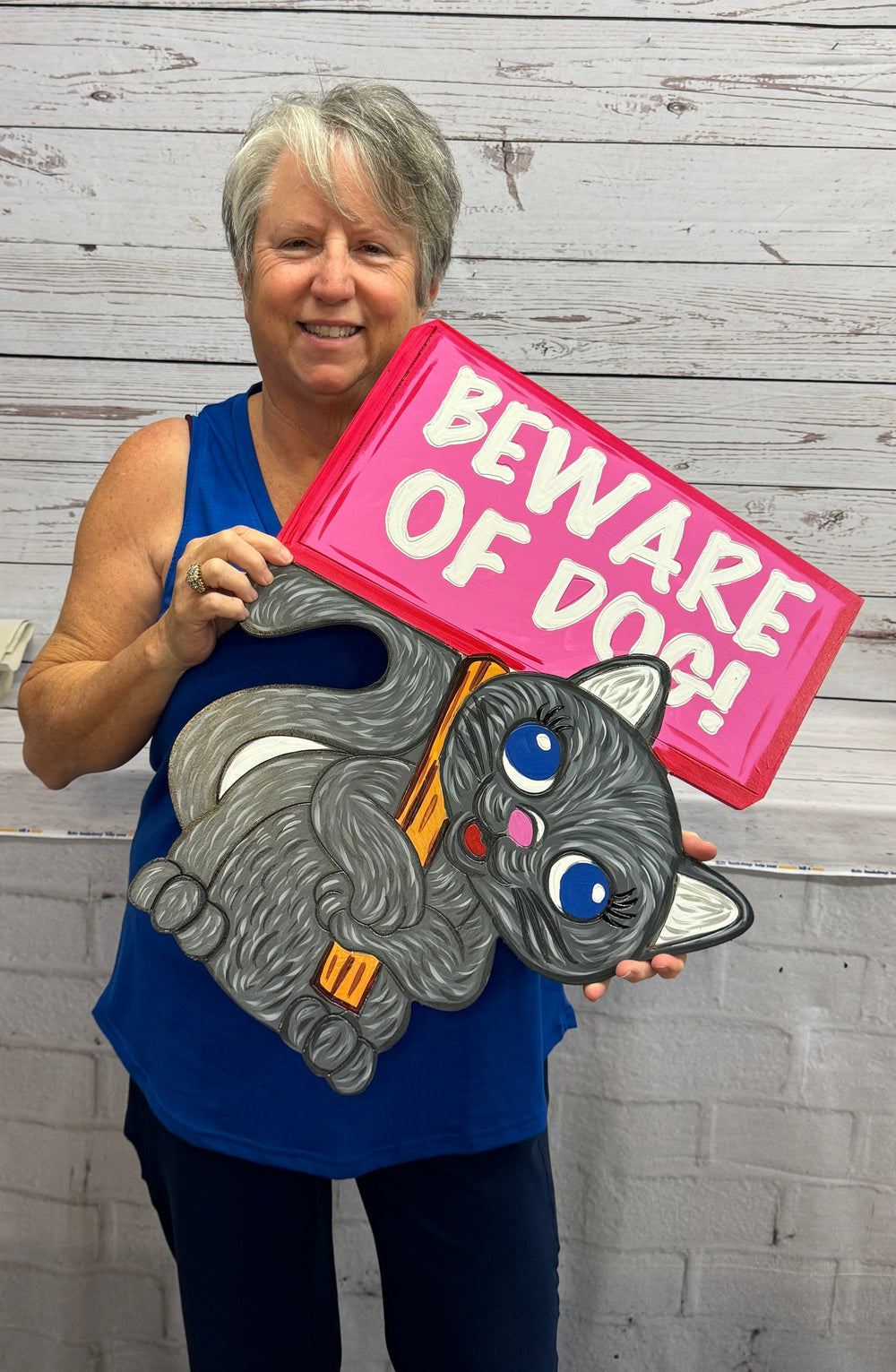 Mary, the owner of Yard Art Decor and More holding a cute, funny decorative animal yard art sign of a gray kitten holding a red "Beware of Dog" sign.  Decorative animal yard signs available for sale at Yard Art Decor and More. 