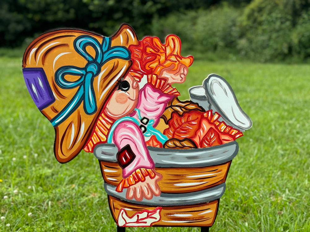 A colorful scarecrow yard sign featuring a scarecrow sitting in a bucket, with autumn-themed details such as a pumpkin and leaves.