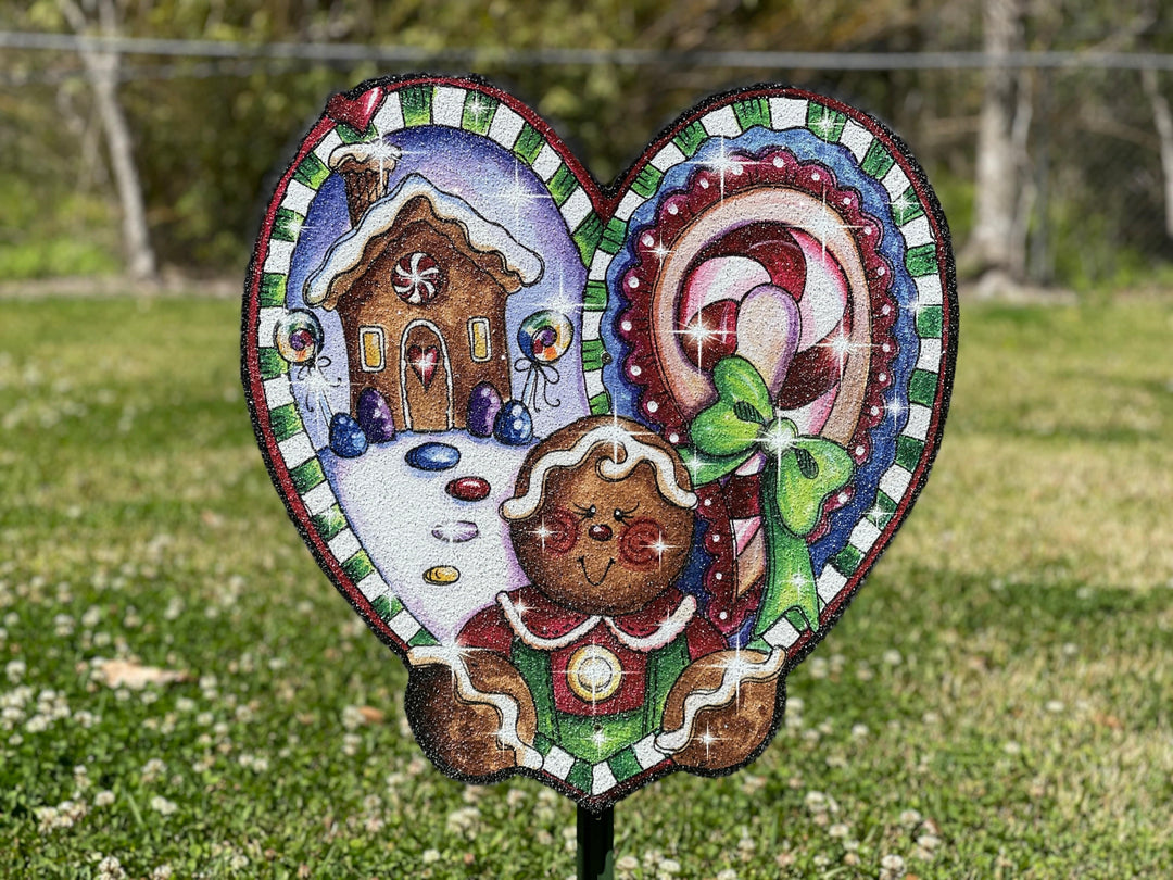 Adorable gingerbread outdoor decorative yard sign in the shape of a heart. Heart has a gingerbread woman on it, along with a picture of a gingerbread house and candy canes. Cute holiday yard signs with stakes for easy install and placement. 