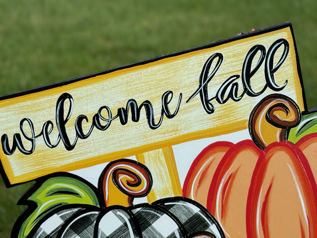 close view of sign on Welcome Fall Sign on top of three Fall pumpkins yard art decoration: one pumpkin is black and white plaid, one pumpkin is leopard print, and one pumpkin is orange