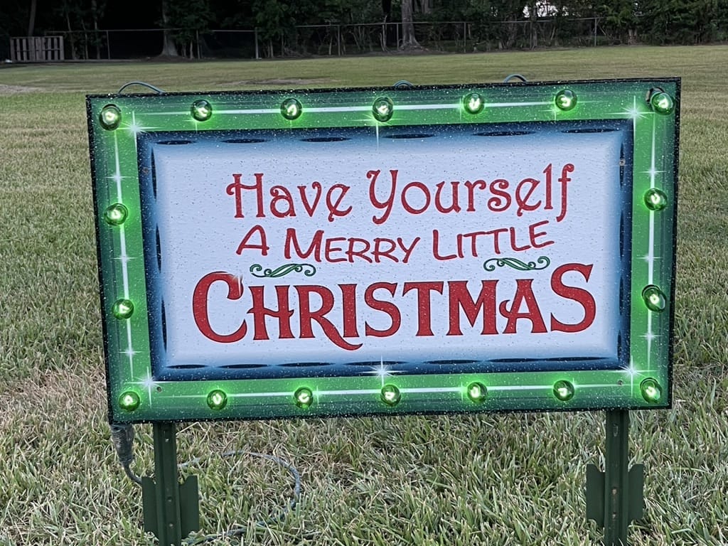 Custom wooden Have Yourself a Merry Little Christmas outdoor yard sign with a hand-painted green border and glowing green C9 incandescent bulbs on a grass lawn.
