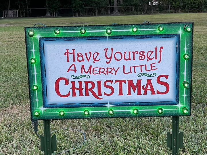 Custom wooden Have Yourself a Merry Little Christmas outdoor yard sign with a hand-painted green border and glowing green C9 incandescent bulbs on a grass lawn.
