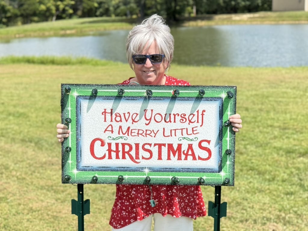 A smiling woman holding a handcrafted, illuminated "Have Yourself a Merry Little Christmas" wooden yard sign featuring a festive green border and built-in glowing lights.