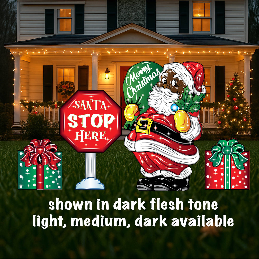 A yard sign featuring a smiling Santa Claus holding a mistletoe and a green bag, accompanied by a 'Santa Stop Here' sign, a lamp post, and presents placed on the grass in front of a house with Christmas decorations.