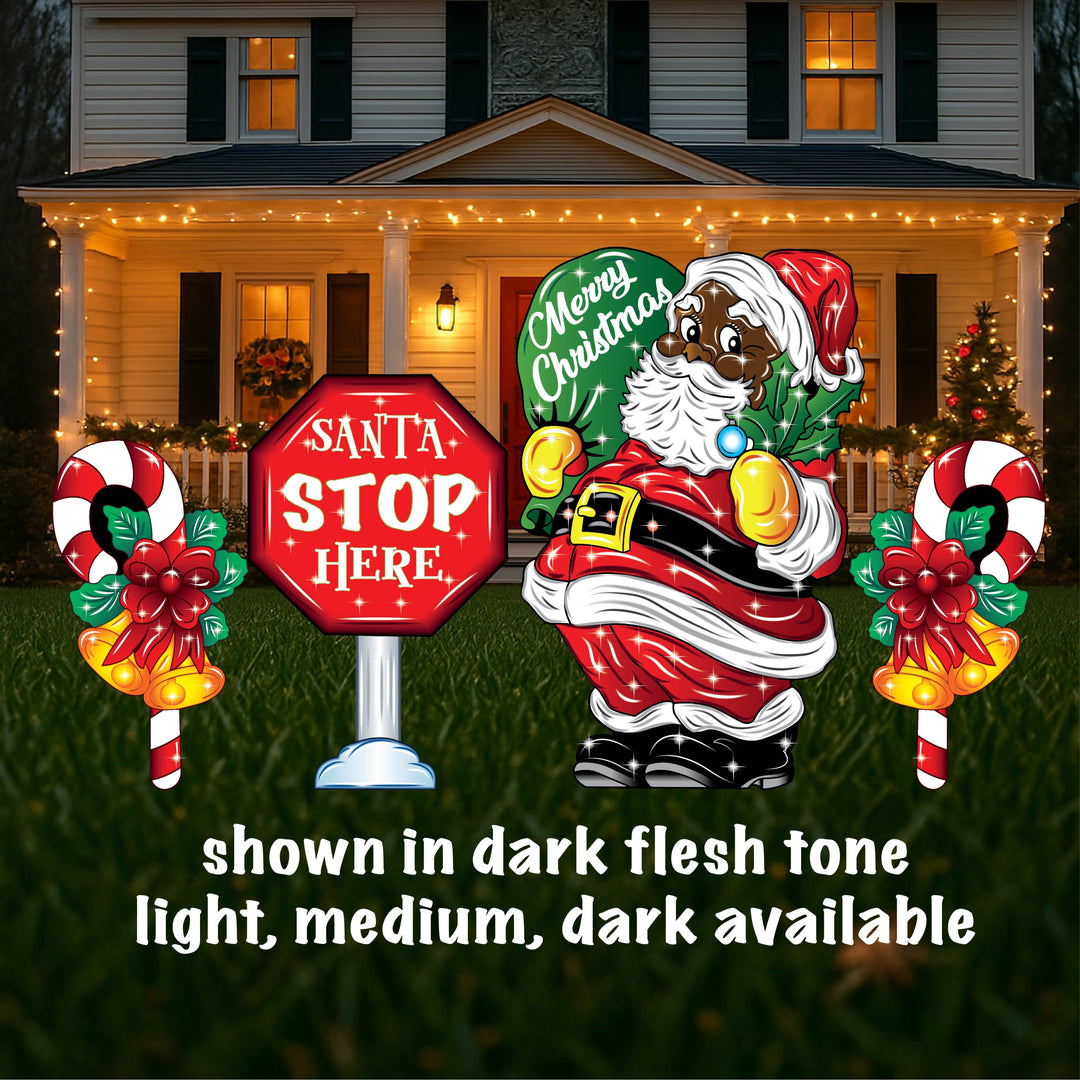 A yard sign featuring a smiling Santa Claus holding a mistletoe and a green bag, accompanied by a 'Santa Stop Here' sign, a lamp post, and presents placed on the grass in front of a house with Christmas decorations.