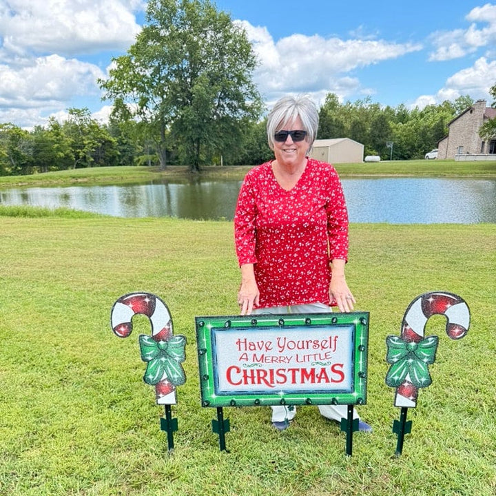 Deluxe outdoor Christmas decoration bundle featuring an illuminated wooden sign and two matching handcrafted candy cane lawn ornaments held on a lawn for size reference.