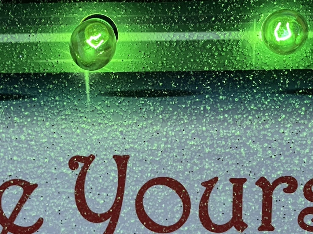 Macro close-up of a customized wooden Christmas display showing red script lettering, a sparkling glitter finish, and vibrant green C9 incandescent bulbs.