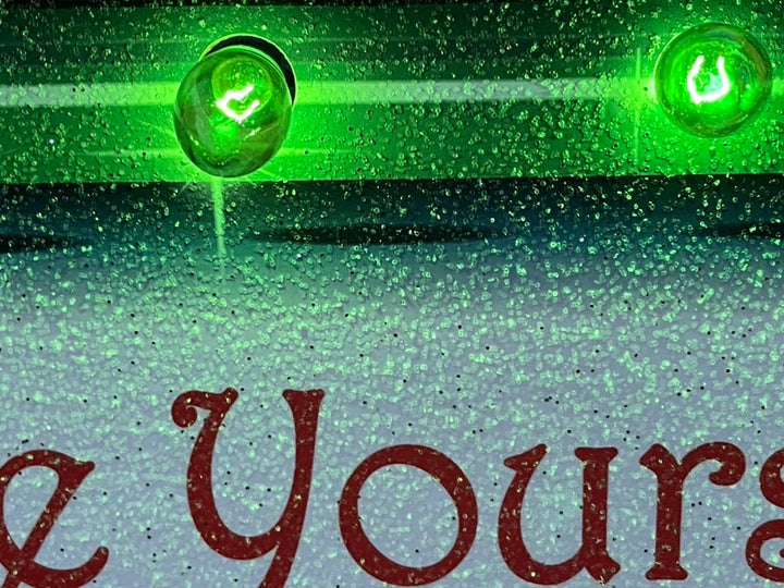 Macro close-up of a customized wooden Christmas display showing red script lettering, a sparkling glitter finish, and vibrant green C9 incandescent bulbs.