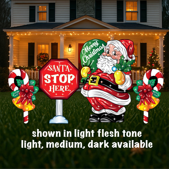 A yard sign featuring a smiling Santa Claus holding a mistletoe and a green bag, accompanied by a 'Santa Stop Here' sign, a lamp post, and presents placed on the grass in front of a house with Christmas decorations.