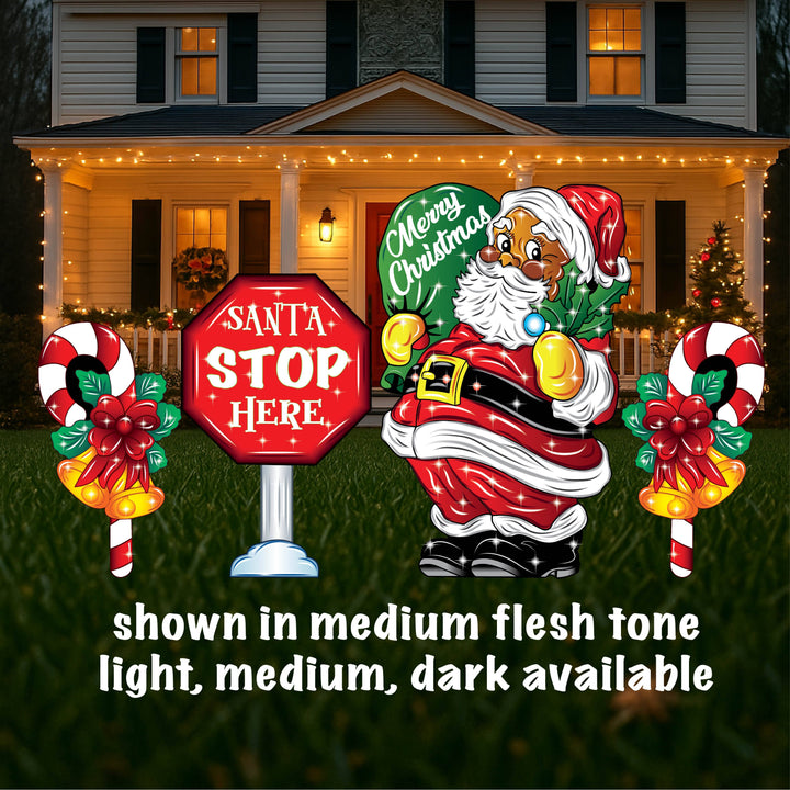 A yard sign featuring a smiling Santa Claus holding a mistletoe and a green bag, accompanied by a 'Santa Stop Here' sign, a lamp post, and presents placed on the grass in front of a house with Christmas decorations.