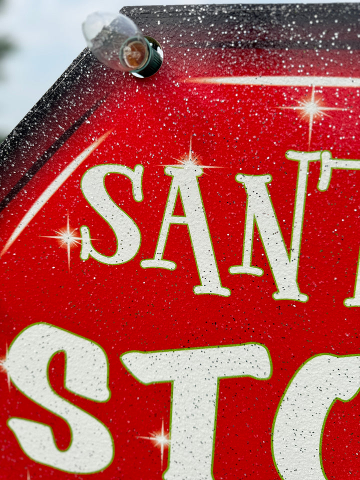 Close-up of red “Santa Stop Here” yard sign with white glitter finish and attached holiday bulb – durable outdoor Christmas decor detail.