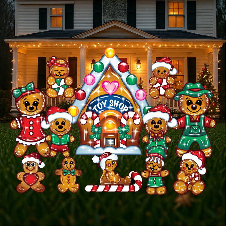 A set of Christmas yard art figures depicting gingerbread characters, including a toy shop, adults, and children, arranged in a display outside a house with holiday decorations.